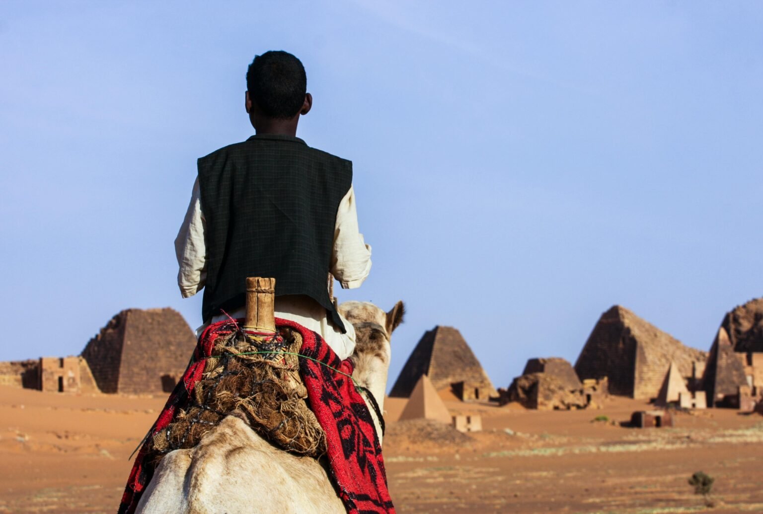 من نحن A man rides a camel towards the historic pyramids of Meroe in the desert of Sudan.